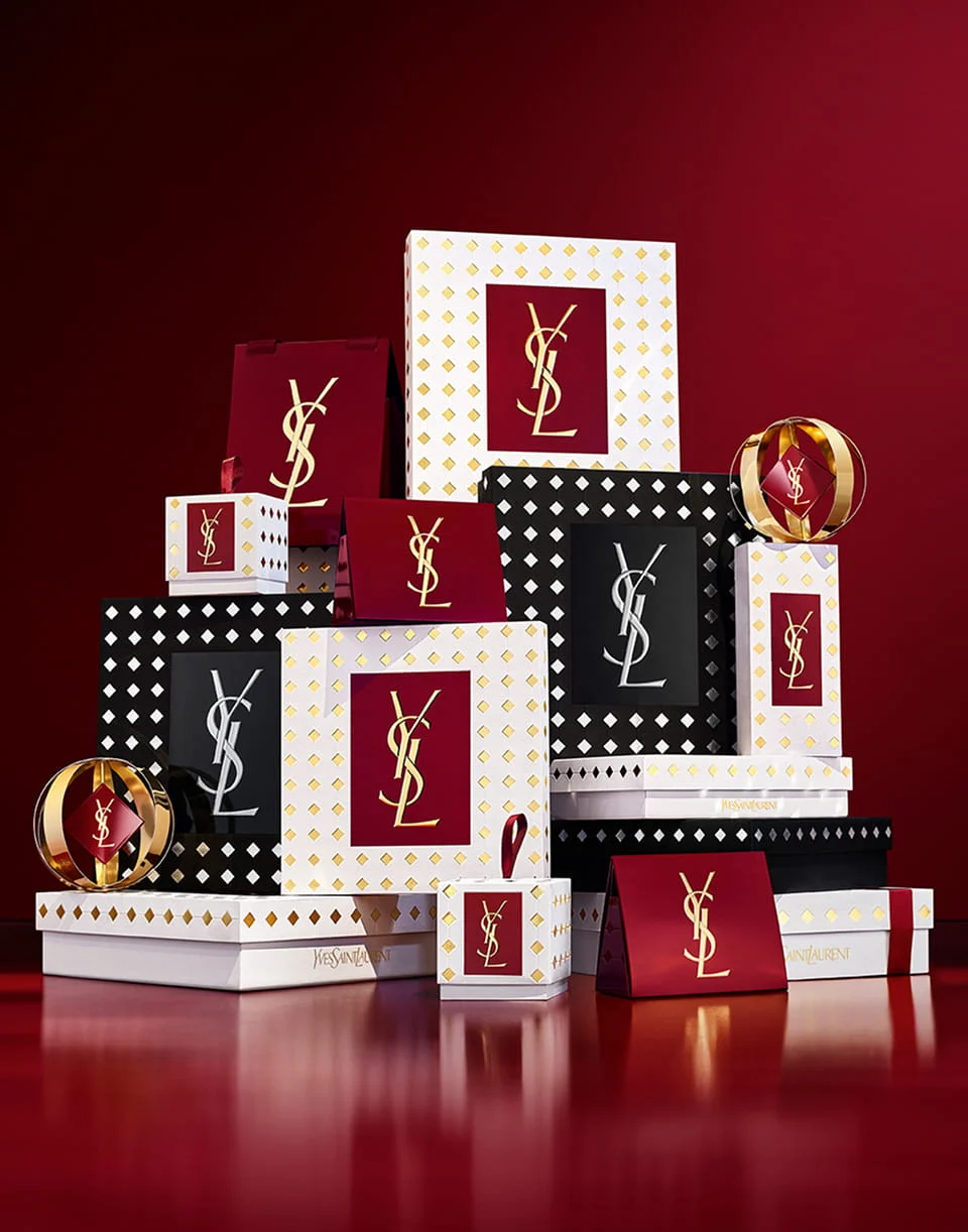 A luxury stack of YSL holiday gift boxes in white, gold, burgundy, and studded black patterns with metallic logos, arranged on a reflective red surface.