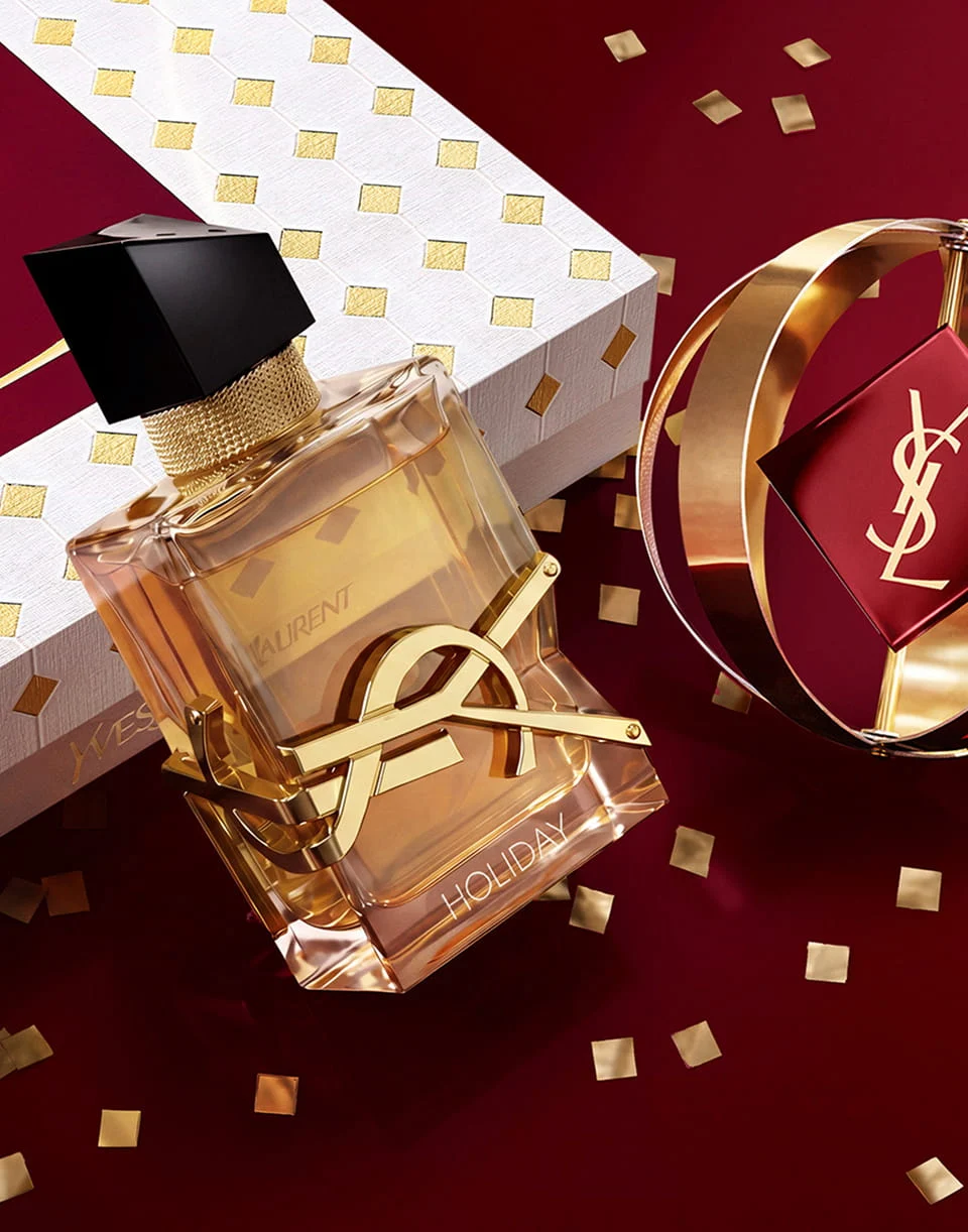 YSL Libre perfume bottle with "HOLIDAY" engraved at the bottom, featuring a bold gold logo resting against a white and gold diamond-patterned gift box with gold confetti on a deep red surface.