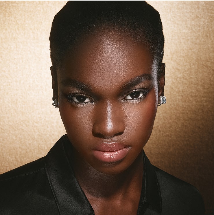 Model with shimmering rose makeup, elegant jewelry, on a gold-toned background.