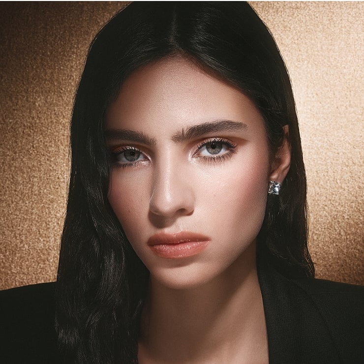 Model Loli Bahia wearing Sparkling Dream makeup look with shimmering eyeshadow and glossy lips on a gold background.