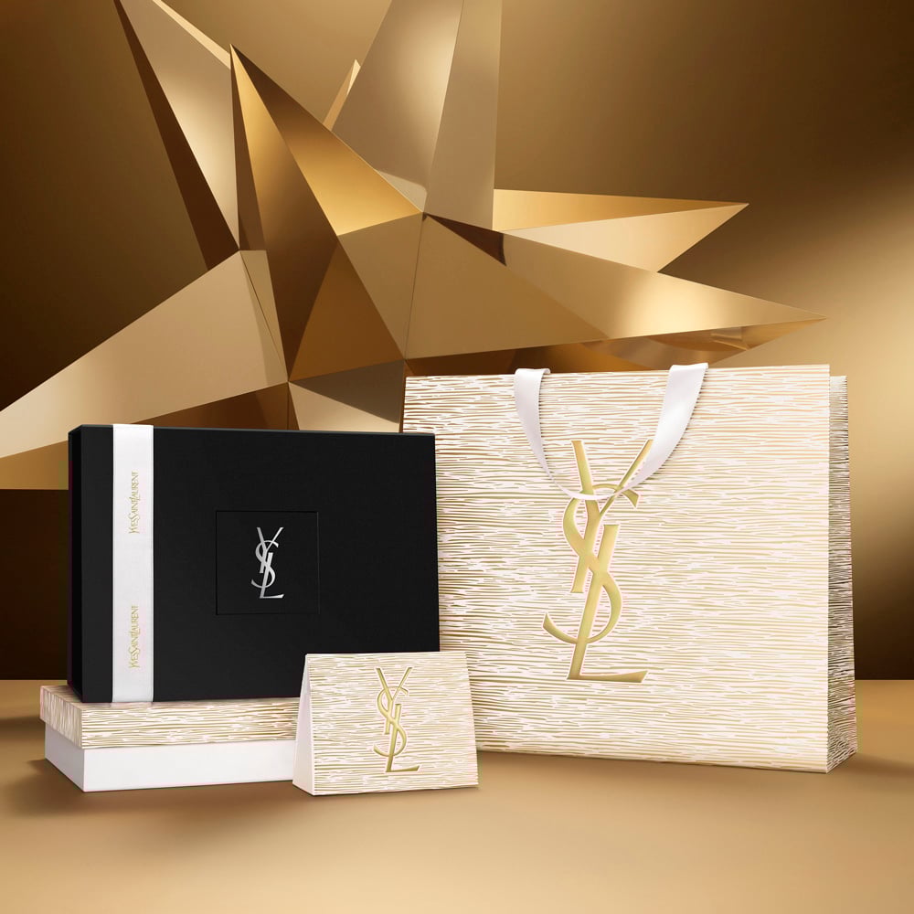Holiday gift set featuring a gold bag with YSL Beauty logo and a black box tied with a white ribbon, displayed on a starry gold-toned background.
