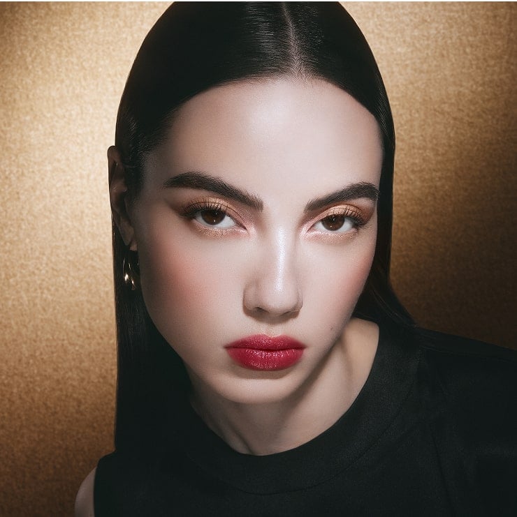 Model with vibrant raspberry makeup, gold jewelry, on a gold-toned background.